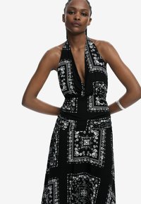 Schwarzes Neckholder-Kleid mit weißen Blumen- und Paisleymustern, mit enganliegendem Oberteil und fließendem Rock. Glattes, leichtes Material.
