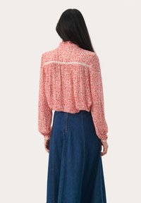 Blusa a maniche lunghe in rosso chiaro con un motivo floreale, dotata di uno schienale arricciato e delicati dettagli in pizzo. Abbinata a una gonna di jeans a vita alta.