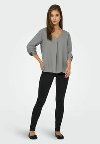 Blusa gris, de corte holgado, con escote en V y mangas enrollables, combinada con vaqueros ajustados negros y bailarinas negras. Textura de tejido suave.