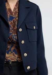 Marineblauwe cropjasje met goudkleurige knopen, gestructureerde stof en een borstzak, gelaagd over een bruin en blauw patroon top.