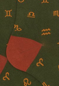 Calzini verdi con segni zodiacali arancioni e un tallone rosso. Il materiale ha una texture liscia e presenta un design senza cuciture.
