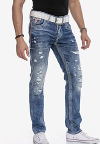 Blaue Jeans aus Denim mit abgenutzten Details, die Risse aufweisen, weiße Nähte und einen gemusterten weißen Gürtel haben. Kombiniert mit weißen Sneakers.