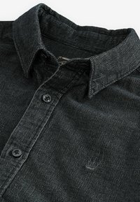 Camisa de pana negra con cuello abotonado, tapeta frontal con botones y bordado de logotipo discreto en el pecho. Tejido texturizado.
