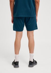 Teal sportieve shorts met een elastische tailleband, gedragen met zwarte sportschoenen en witte sokken met zwarte patronen, vanaf de achterkant bekeken.