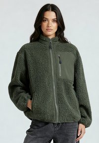 Veste en polaire vert olive avec un panneau avant lisse, un col montant, une poche poitrine zippée, un tissu texturé et des épaules tombantes.