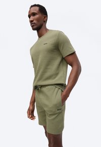 Mees, kes kannab oliivirohelisi lühikese varrukaga särki ja sobivaid lühikesi pükse, millel on rinnal ja reiel BOSSi logo, seisab ühe käega taskus.