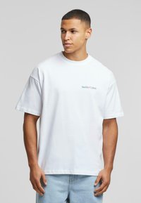 Bílý bavlněný tričko s uvolněným střihem, krátkými rukávy a malým modrým logem "Another Cotton" na levé straně hrudi.