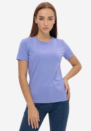 Giovane donna con capelli castani lisci indossa una maglietta viola chiaro a maniche corte e jeans blu scuro, in piedi con una mano sul fianco.