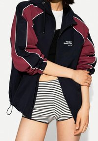 Person wearing a black and maroon zip-up jacket with "Nautical Sports Club" logo, black top, and black-and-white striped shorts, arms crossed.
