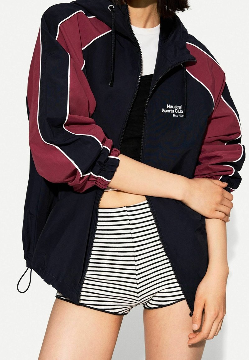 Person wearing a black and maroon zip-up jacket with "Nautical Sports Club" logo, black top, and black-and-white striped shorts, arms crossed.