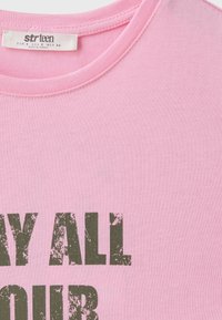 T-shirt rose en coton avec col rond et une étiquette indiquant la taille et l'origine, texte partiellement visible en police verte foncée usée.