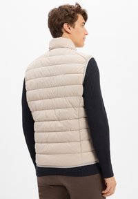Junger Mann, der eine beige gesteppte Weste über einem schwarzen gerippten Langarmpullover und braunen Hosen trägt, seitlich blickend vor einem neutralen Hintergrund.