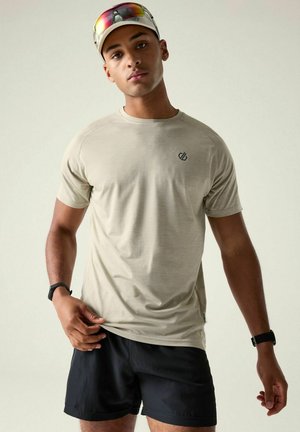 Jonge man draagt een beige T-shirt, zwarte korte broek, een lichte pet met reflecterende zonnebril en een zwart polshorloge aan elke pols.