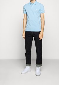 Man i ljusblå pikétröja, mörka upprullade jeans och vita högstövlar står mot en enkel bakgrund.