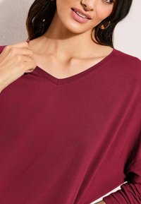 Burgundy V-neck top with long sleeves, made from soft fabric, featuring a relaxed fit and a smooth texture. Minimal seams accentuate design.