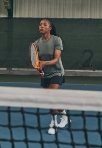 Pelēka sporta krekls un melni šorti, ko nēsā spēlētājs, kurš tur oranžu tenisa raketi, novietojoties tuvu tīklam zilā kortā.