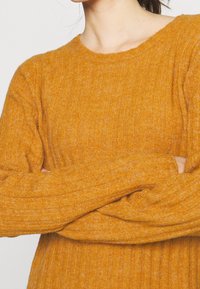 Pull en maille côtelée de couleur moutarde avec des manches longues et un col rond, fabriqué à partir d'un matériau tricoté texturé et doux.
