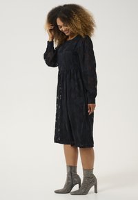 Schwarzes Blumenspitzenkleid mit langen Ärmeln, gerafftem Bund und A-Linien-Silhouette, kombiniert mit glitzernden silbernen Knöchelstiefeln.