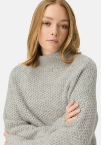 Szary sweter z dzianiny waffle, z wysokim kołnierzem, długimi rękawami i luźnym krojem, wykonany z fakturowanej tkaniny. Szczegóły obejmują ściągacze przy mankietach.
