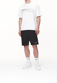 Mężczyzna w białej, przew oversize t-shircie z napisem "PEGADOR", czarnych szortach, białych skarpetkach i białych sneakersach stoi na tle białego tła.