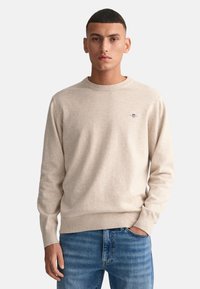 Beige gestrickter Pullover mit Rundhalsausschnitt und langen Ärmeln, der ein kleines gesticktes Logo auf der Brust sowie gerippte Bündchen und Saum aufweist.