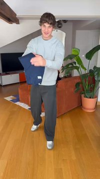 Conjunto casual com um suéter cinza claro e calças largas azul-marinho. A pessoa está segurando um item dobrado. Ambiente interno com plantas.