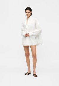 Γυναίκα που στέκεται φορώντας ένα υπερμεγέθες λευκό πουκάμισο με κουμπιά, μαύρα σανδάλια και λιτό μακιγιάζ σε απλό λευκό φόντο.