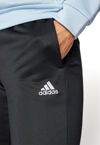 Čierne športové nohavice s hladkým povrchom, vybavené bielym logom Adidas na ľavom stehne a bočnými vreckami. Materiál je ľahký a pružný.
