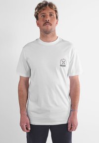 T-shirt en coton blanc avec un col rond et des manches courtes. Le côté gauche de la poitrine comporte un texte graphique noir et des éléments de design.
