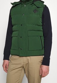 Vihreä toppaliivi, jossa korkea kaulus, mustat napeilla varustetut kiinnitykset, kaksi etutaskua ja timantin muotoinen logolapuke. Päällä tumma paita ja beiget housut.