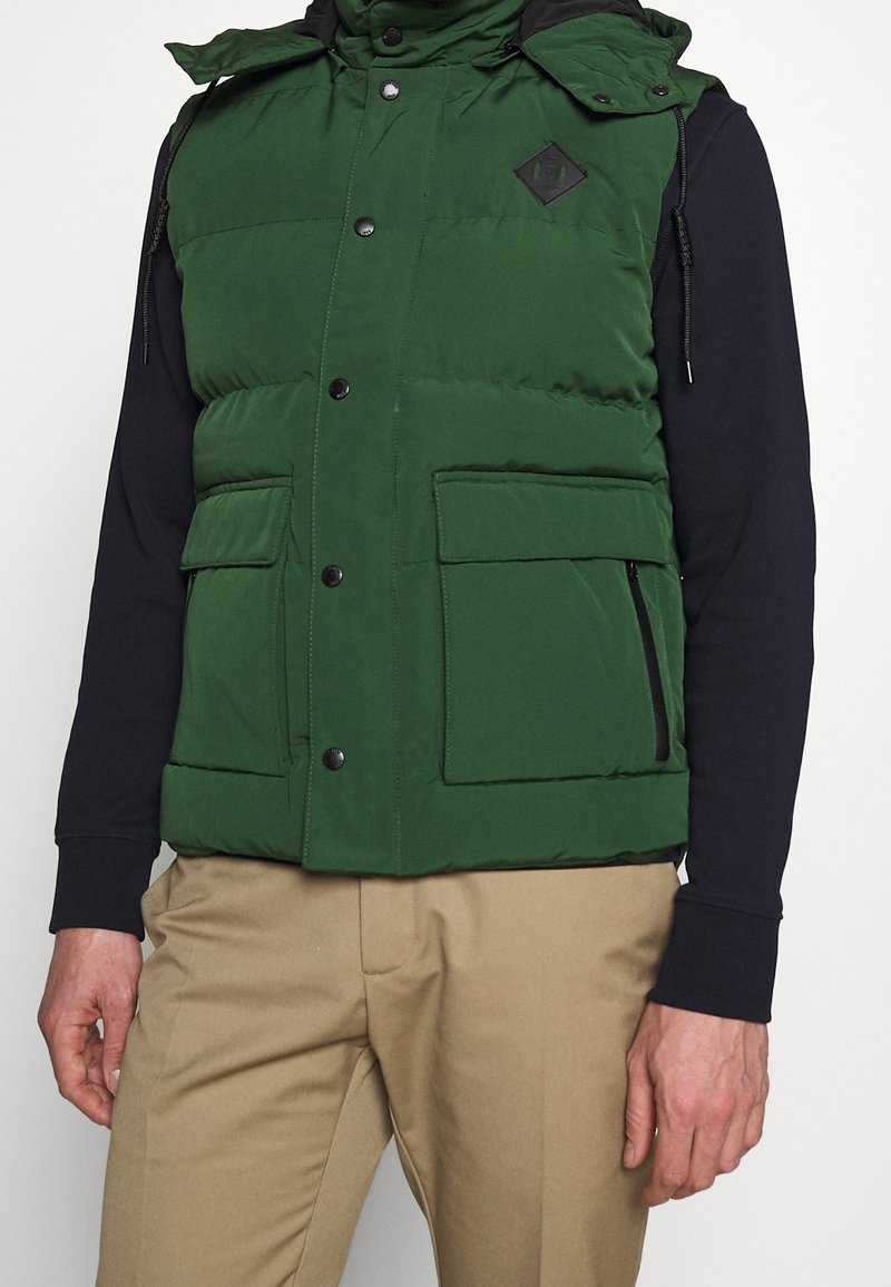 Vihreä toppaliivi, jossa korkea kaulus, mustat napeilla varustetut kiinnitykset, kaksi etutaskua ja timantin muotoinen logolapuke. Päällä tumma paita ja beiget housut.