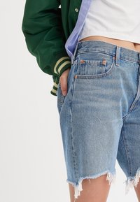 Pantalones cortos de mezclilla en azul claro con dobladillo deshilachado, adornados con detalles metálicos en cobre y un corte relajado, combinados con una chaqueta bomber verde.