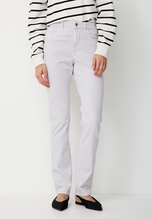 Person trägt hellgraue gerade geschnittene Hose, schwarze flache Schuhe und ein langärmeliges weißes Hemd mit schwarzen horizontalen Streifen.