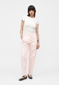 Vrouw met donker haar tot op de schouders draagt een witte nauwsluitende korte mouw top, lichtroze hoge taille broek en zwarte platte schoenen met zilveren studs.