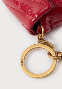 Canto de uma bolsa de couro acolchoado vermelho com um anel redondo de metal dourado preso, apresentando uma inscrição da marca. Textura suave com costuras visíveis.