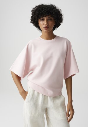 Femme aux cheveux noirs bouclés portant un haut ample rose clair à manches courtes et un pantalon blanc, debout, une main dans la poche.