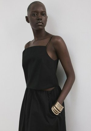 Vrouw met een donkere huid, gekleed in een zwarte mouwloze top en rok, staand tegen een witte muur, met meerdere gouden armbanden om haar pols.