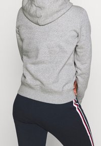 Person som bär en grå huvtröja och svarta byxor med röda, vita och blå sidostråkar, står med ena handen bakom ryggen.