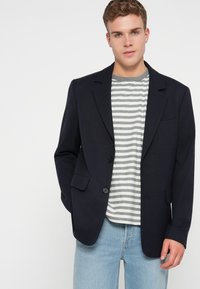 Blazer blu in tessuto testurizzato con rever a notches, chiusura con un bottone e due tasche frontali, indossato su una camicia a righe grigie e bianche.