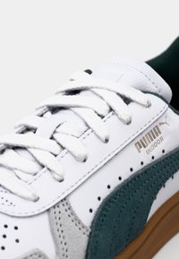 Hvite lær sneakers med grønne og grå semskede detaljer. Har hvite snøringer, perforerte detaljer og en brun gummisåle. Puma-logoen er til stede.