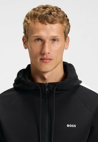Czarna bluza z kapturem na zamek błyskawiczny, z regulowanym kapturem, z białymi akcentami na ramionach oraz małym logo "BOSS" na klatce piersiowej. Gładka faktura tkaniny.