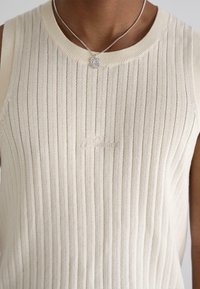 Ribbed, sleeveless cream top with a round neckline and subtle textured pattern. Features delicate embroidered logo on the front.