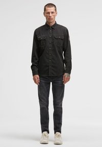 Mann mit neutralem Ausdruck, der ein schwarzes Jeanshemd, dunkle Jeans, weiße Turnschuhe trägt und sichtbare Tattoos am Hals und an den Händen hat.