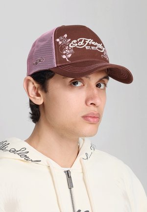 Jeune homme portant une casquette en mesh Ed Hardy marron et rose et un sweat à capuche zippé blanc avec un lettrage noir sur le col.