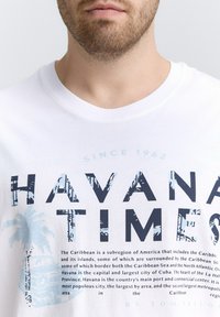 Camiseta de algodón blanca con gráfico "Havana Times" en azul marino y azul claro, con diseño de palmera y disposición del texto, cuello redondo.