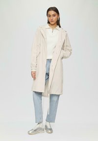 Jasnobeżowy płaszcz przeciwdeszczowy z kapturem, z zatrzaskami i elastycznymi mankietami, noszony na białej bluzce i jasnoniebieskich dżinsach, z srebrnymi sneakersami.