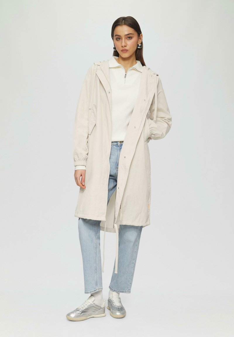 Jasnobeżowy płaszcz przeciwdeszczowy z kapturem, z zatrzaskami i elastycznymi mankietami, noszony na białej bluzce i jasnoniebieskich dżinsach, z srebrnymi sneakersami.