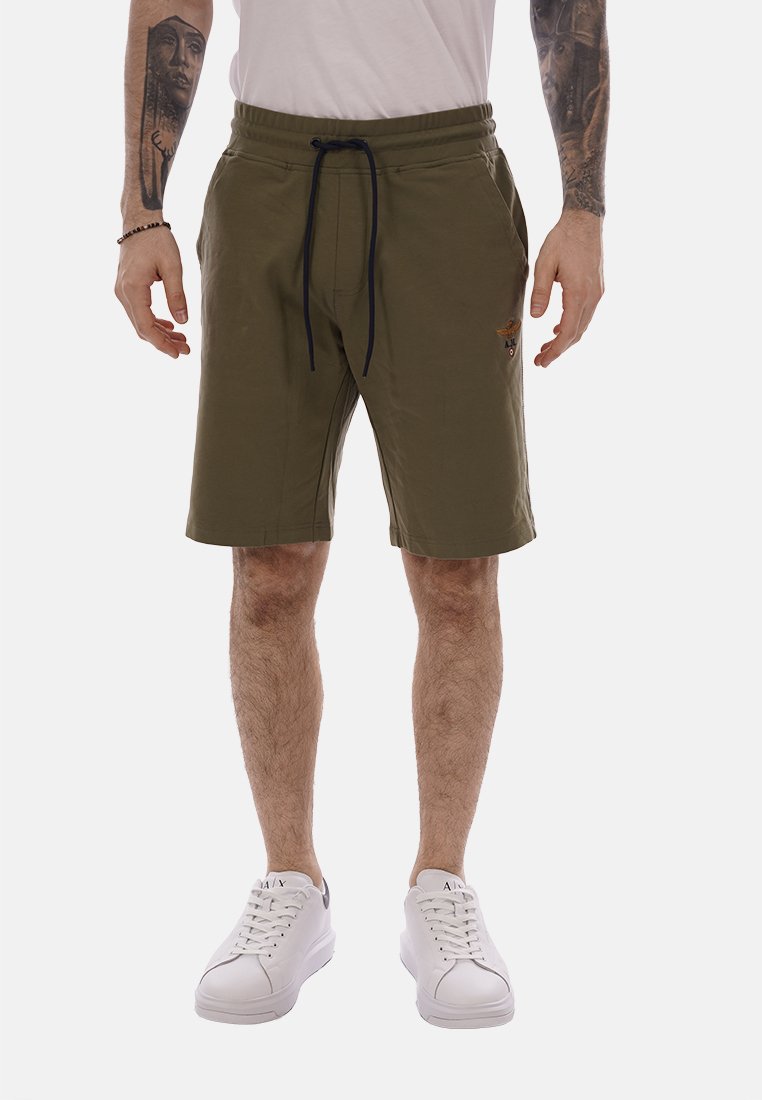 Homme portant un short à cordon de serrage vert olive, des baskets blanches et une chemise blanche, debout avec les bras détendus et les avant-bras tatoués visibles.