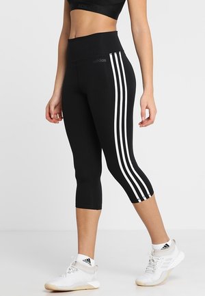Person, die schwarze Adidas Cropped-Leggings mit weißen Streifen und weiße Adidas-Sneaker trägt, steht vor einem schlichten Hintergrund.