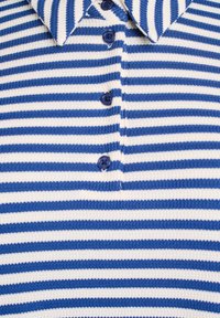 Stickad polo i blå och vita horisontella ränder. Har en spetsig krage och fyra blå knappar framtill. Strukturerad tyg.